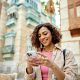 Waist-up view of casually dressed woman with curly brown hair looking at smart phone and smiling while visiting historic Al-Balad, Jeddah.